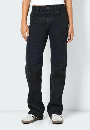 Personne portant un jean noir droit et des baskets blanches, debout devant un fond blanc avec les bras détendus le long du corps.