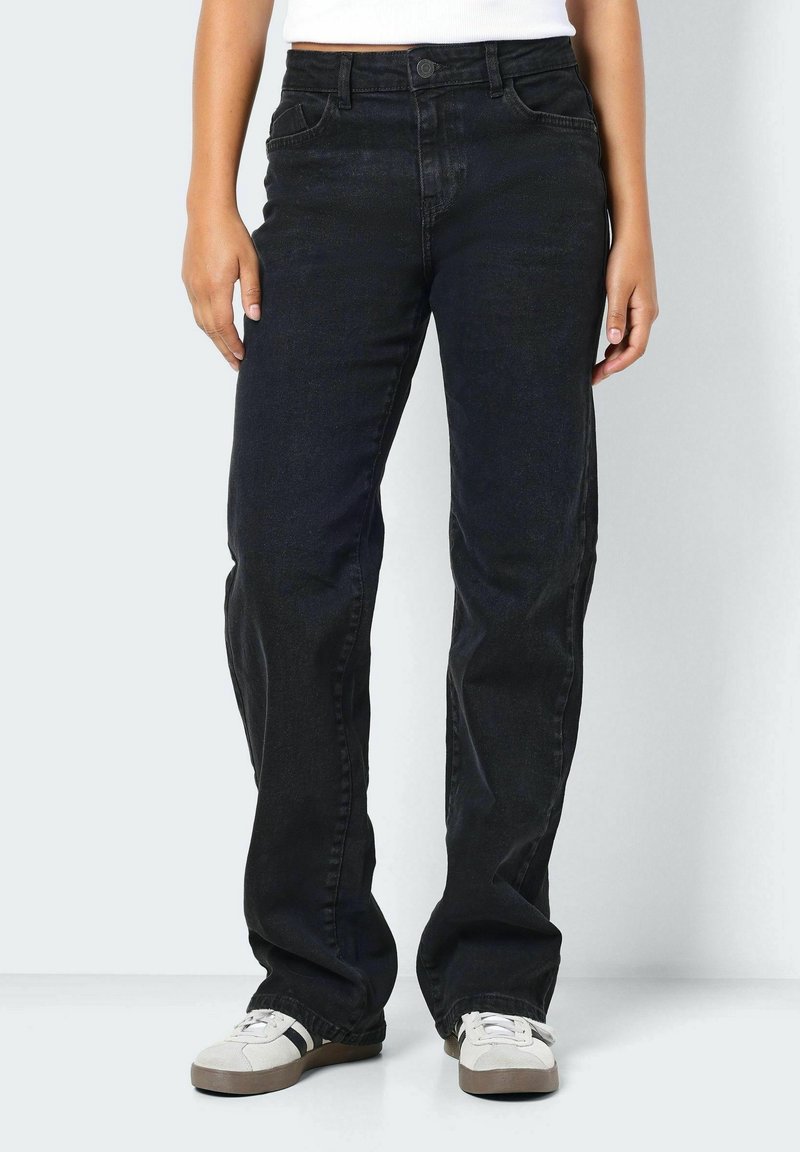 Personne portant un jean noir droit et des baskets blanches, debout devant un fond blanc avec les bras détendus le long du corps.