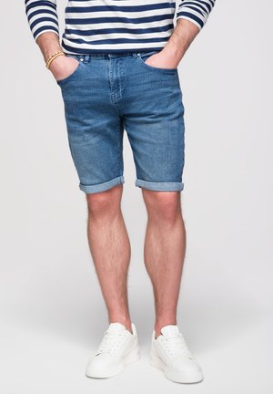 Homme portant un short en jean bleu roulé, des baskets blanches et une chemise rayée à manches longues, mains dans les poches, sur un fond uni.
