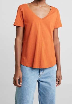 Person trägt ein orangefarbenes T-Shirt mit kurzen Ärmeln und V-Ausschnitt sowie hellblaue Jeans vor einem schlichten Hintergrund.
