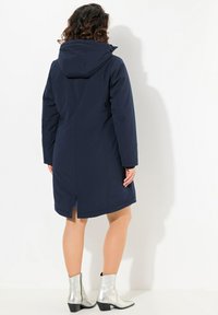 Navy blue waterproof coat with a hood, featuring long sleeves, a straight cut, and a back vent. Worn with silver ankle boots.