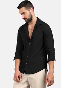 Maglietta nera a maniche lunghe in tessuto testurizzato, con un motivo discreto, chiusura con bottone e colletto a punte, abbinata a pantaloni beige.