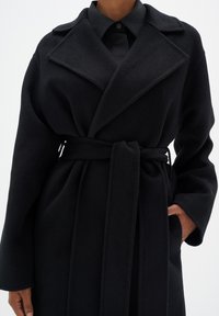 Manteau noir en tissu doux et texturé avec une taille ceinturée, de grands revers et des poches latérales visibles, conçu pour un ajustement décontracté.