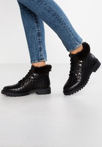 Botines negros acolchados con borde de piel sintética, diseño con cordones, detalles de herrajes metálicos y suela de goma texturizada, combinados con vaqueros azules.