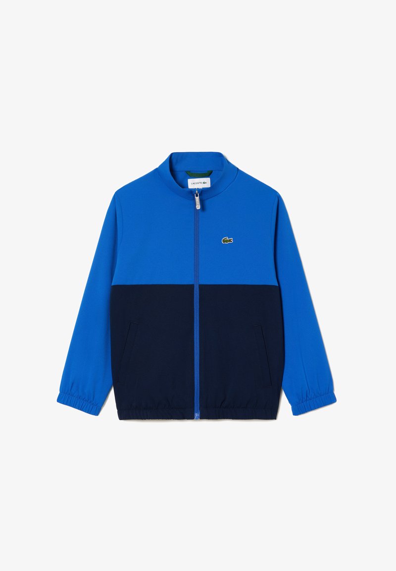 Veste zippée bleu et marine avec col montant, poignets élastiques et poches latérales, arborant un petit logo vert sur la poitrine.