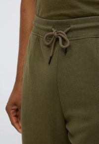 Ελιά πράσινο παντελόνι φόρμας με εσώτατη ζώνη σύσφιξης, κατασκευασμένο από παχύ υφή υφάσματος και μαύρα μεταλλικά άγκιστρα στη ζώνη σύσφιξης.