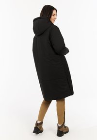 Abrigo inflado negro con capucha, de diseño largo, con paneles cosidos y bolsillos laterales. Combinado con mallas color tostado y botas marrones.