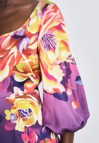 Das Blumenmusterkleid weist lebendige Farben mit großen pinken, gelben und lila Blüten auf. Der Stoff ist glatt und hat ein puffiges Ärmel-Design.