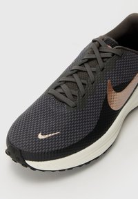 Běžecká obuv Nike s šedým síťovaným svrškem, černými akcenty a růžovo-zlatým logem Swoosh. Obsahuje texturované plochy a polstrovanou podrážku.