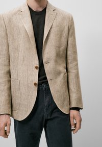 Beżowa marynarka w jodełkę z dwoma guzikami, klapami z wycięciem i kieszonką na piersi, noszona na ciemnym swetrze i czarnych spodniach.