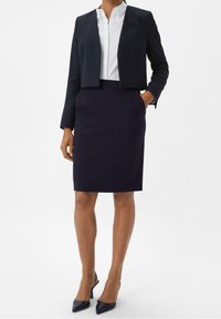 Marineblazer mit kurzem Schnitt über einem weißen Hemd mit Kragen, kombiniert mit einem taillierten marineblauen Bleistiftrock und spitzen schwarzen Absatzschuhen.