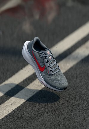 Grå Nike løbesko med rødt swoosh på asfalt nær hvid vejlinje, vist i luften med små stykker omkring den.