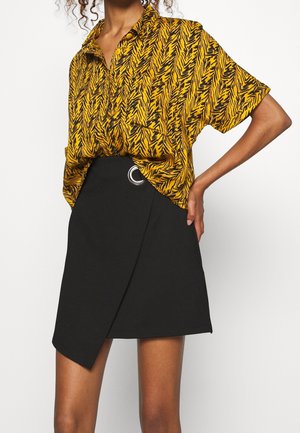 Kvinde iført en gul og sort mønstret kortærmet skjorte indtullet i et sort asymmetrisk skørt med en sølv ringdetalje.