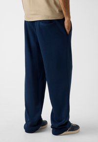 Marineblaue Sweathosen mit lockerem Schnitt, Seitentaschen und weicher Textur. Getragen mit dunklen Schuhen, die ein beiges und marineblaues Farbschema zeigen.