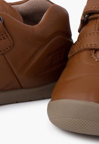 Scarpe in pelle marrone con una texture liscia, dotate di una fascia in Velcro e design a punta arrotondata. Suola in gomma con una presa a motivo.