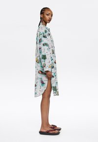 Abito camicia leggero e con maniche lunghe in tessuto trasparente, caratterizzato da una stampa colorata a tema natura. Presenta un orlo curvo e fessure laterali. Sandali neri.