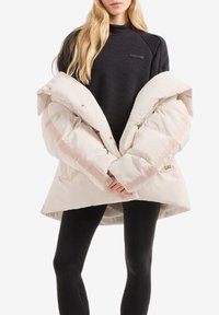 Giacca a vento in morbido beige con dettagli rosa, caratterizzata da un grande colletto, vita stretta e tasche laterali, indossata sopra un top nero e leggings.