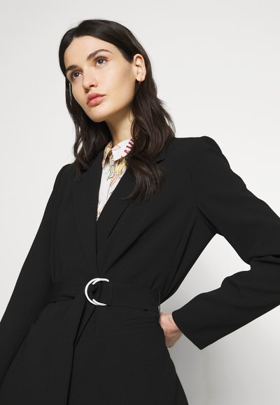 Cappotto nero su misura con vita cinte e fibbia argentata. Presenta maniche lunghe, colletto a risvolto e tasche laterali. Tessuto liscio e strutturato.