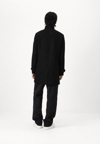 Abrigo de lana negro con cuello alto, corte recto y abertura trasera. Combinado con pantalones negros holgados y zapatillas blancas, visto desde atrás.