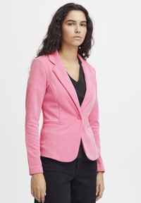 Blazer rosa in tessuto morbido, design aderente con rever a punta, chiusura con un solo bottone e tasche frontali. Indossato sopra un top nero a scollo a V.