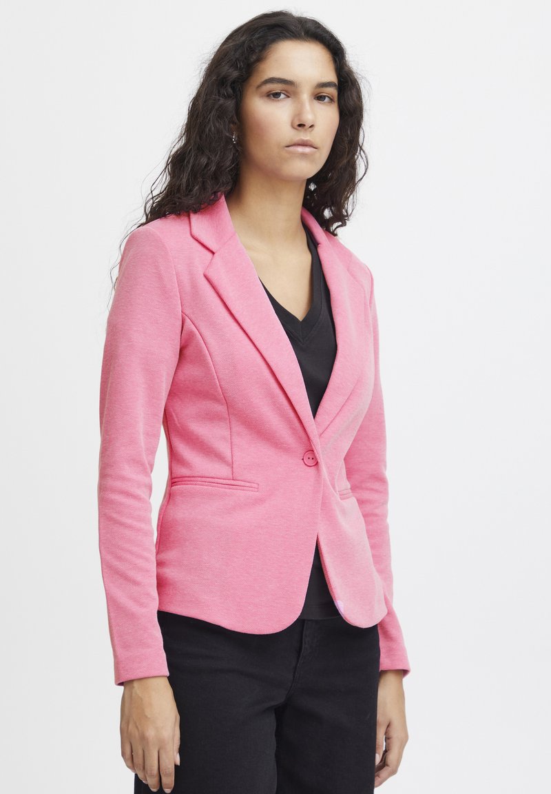 Blazer rosa in tessuto morbido, design aderente con rever a punta, chiusura con un solo bottone e tasche frontali. Indossato sopra un top nero a scollo a V.