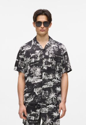 Sort kortærmet skjorte med hvidt tropisk mønster, der forestiller palmer og landskaber. Kombineret med sorte solbriller.