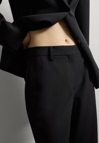 Pantalons noirs sur mesure avec une texture lisse, dotés d'une fermeture éclair et d'un bouton, de poches latérales et d'un design à pli plat. Coupe slim.