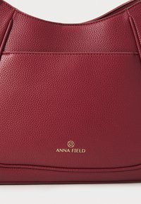 Sac à épaule en cuir bourgogne avec une texture granuleuse, présentant un logo doré sur le bas à l'avant et une silhouette courbée et élégante.