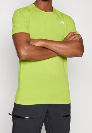 Mies, jolla on kirkkaanvihreä lyhythihainen The North Face -t-paita, kädet ristissä, yhdistettynä mustiin housuihin, joissa on vaakasuorat vetoketjutaskut.