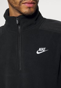 Schwarzer Fleece-Pullover mit Viertelreißverschluss und hohem Kragen, der ein weißes Nike-Logo auf der Brust und strukturierte Schulterakzente aufweist.