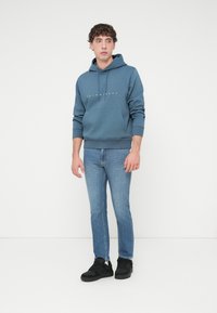 Μπλε φούτερ με κουκούλα και μπροστινή τσέπη, με λευκό κείμενο λογότυπου, συνδυασμένο με ανοιχτόχρωμα τζιν και μαύρα αθλητικά παπούτσια. Λεπτή υφή υφάσματος.