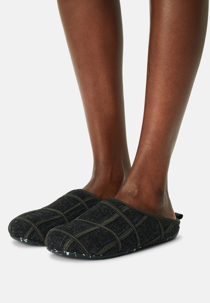 Pieds portant des chaussons gris foncé à enfiler avec des motifs croisés cousus et des semelles texturées sur un fond clair uni.