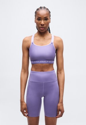 Vrouw met gevlochten haar, gekleed in een bijpassende lavendelkleurige sportbeha en hoog getailleerde fietsbroek, staand tegen een effen achtergrond.
