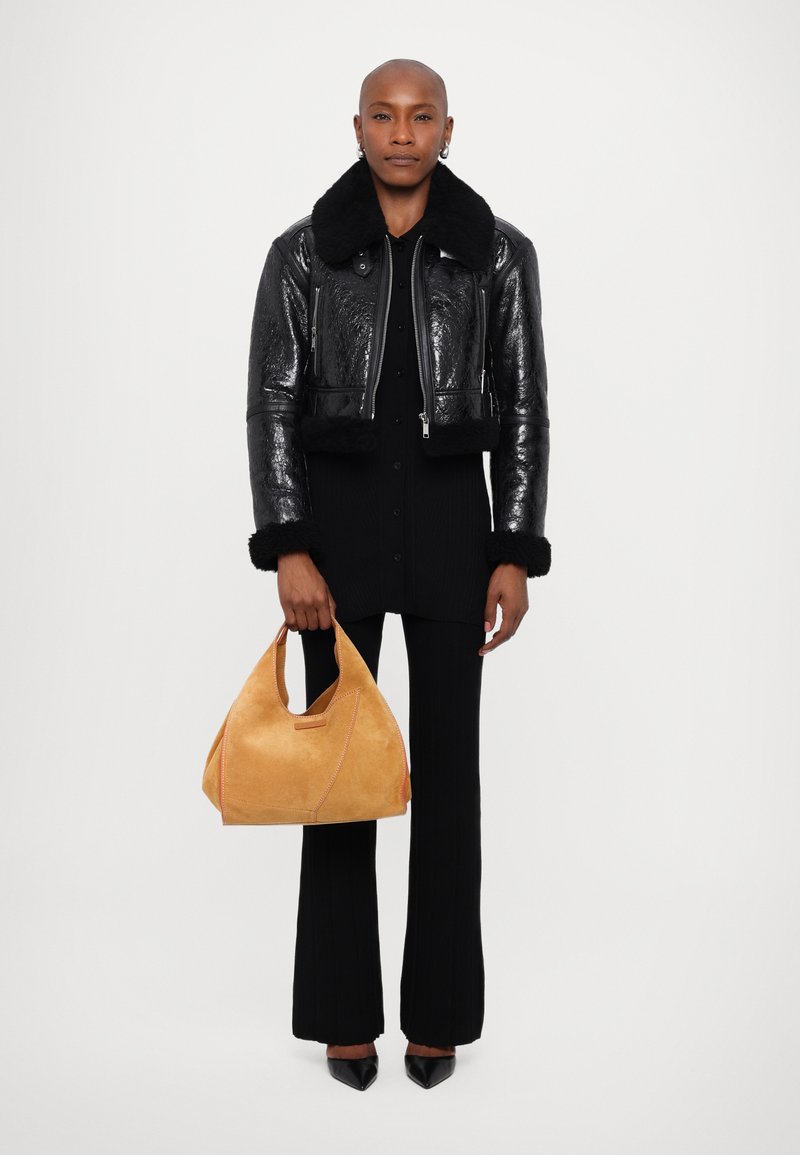 Veste bomber en cuir noir avec col en fausse fourrure, associée à un top noir côtelé et un pantalon noir évasé. Tenant un sac à main en suède jaune avec des accents.