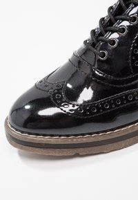Chaussure brogue en cuir noir brillant avec des détails perforés, devant à lacets et semelle épaisse en caoutchouc marron.
