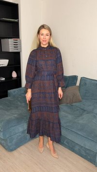 Vrouw binnenstaand in een midi-jurk met lange mouwen en patroon, beige hakken, met een kleine bruine clutch naast een blauwe bank.