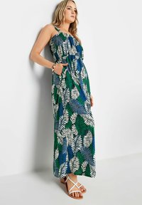 Maxi dress realizzato in tessuto con motivi a foglie verdi, blu e bianchi; design senza maniche con tasche laterali; abbinato a sandali bianchi.