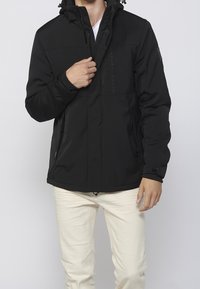 Veste noire isolante avec fermeture éclair à l'avant, capuche et deux poches latérales. Caractérisée par un matériau texturé et un logo sur la poitrine.