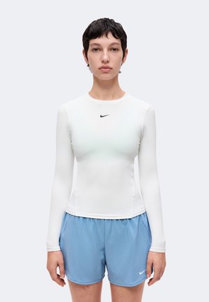 Persoon met kort donker haar, gekleed in een aansluitend wit shirt met lange mouwen en lichtblauwe sportshorts met Nike-logo's, staand tegen een effen achtergrond.