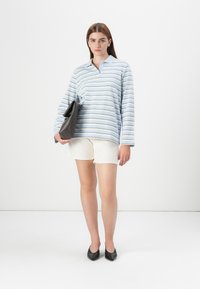 Model stands in a white background, wearing a blue striped oversized shirt, white shorts, and black heels, holding a dark handbag.