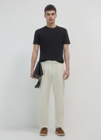 Camicia nera a maniche corte, pantaloni color crema, scarpe di camoscio marrone. Tiene una borsa di pelle nera con finitura liscia e design elegante.