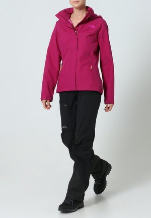 Personne portant une veste à capuche magenta, un pantalon noir et des chaussures de randonnée noires, marchant sur un fond clair uni.