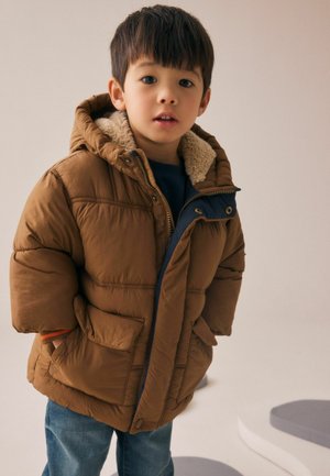 Giacca a vento marrone con cappuccio foderato in pile, dettagli blu navy, zip frontale e due tasche laterali, indossata sopra un maglione blu e jeans.