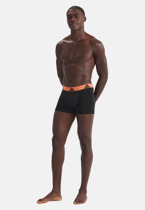 Athletischer Mann steht barfuß und trägt schwarze Boxershorts mit einem orangen Bund, auf dem ein schwarzes Adidas-Logo zu sehen ist, vor einem schlichten weißen Hintergrund.