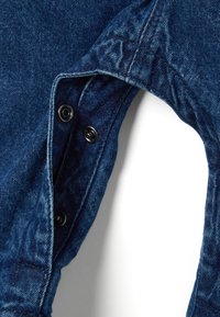 Tela de denim en azul oscuro, con una abertura curva y botones a presión negros. La textura está ligeramente desvanecida con detalles de costura visibles.