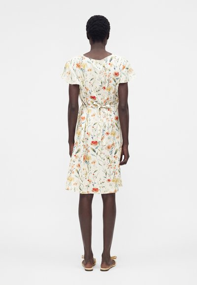 Γυναίκα που φορά λευκό φόρεμα με λουλούδια μέχρι το γόνατο, κοντά μανίκια και δεμένο ζώνη, στέκεται με την πλάτη σε απλό φόντο.