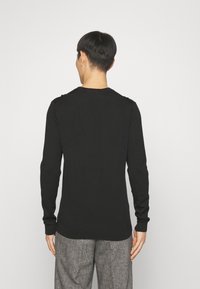 Homme se tenant de dos, portant un pull noir à manches longues et un pantalon gris texturé sur un fond clair et uni.