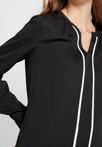 Svart blus med djup V-ringning och vit kantning nedför framsidan, med långa ärmar och en slät, glänsande yta.