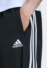 Čierne športové nohavice s bielymi pruhmi, obsahujúce zipsovú kapsu a vyšitým bielym logom Adidas na hornej ľavej stehne.