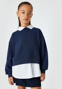 Marinefarbener Oversized-Pullover mit einer weißen, kragenbasierten Schicht darunter, die gerippte Bündchen und Saum sowie hochgekrempelte Ärmel für einen lässigen Sitz aufweist.